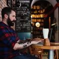 Bild in Galerie-Betrachter laden, En skægget mand med tatoveringer, der ligner en moderne viking, er iført en rød og blå plaid skjorte, mens han taster på en bærbar computer ved et rundt træbord i en hyggelig café med bogreoler og tavlevægge. En ged Kaffe Drikkehorn Brun isoleret kop lavet af BPA-fri plastik sidder tæt på hans bærbare computer.