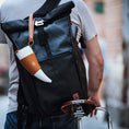 Bild in Galerie-Betrachter laden, En person iført en grå t-shirt bærer en stor sort roll-top rygsæk med en hvid og solbrun ged Kaffe Drikkehorn Brun påsat, mens han skubber en cykel med en brun Brooks-sadel i læder. Scenen fremkalder stemningen fra en moderne viking på en bygade.