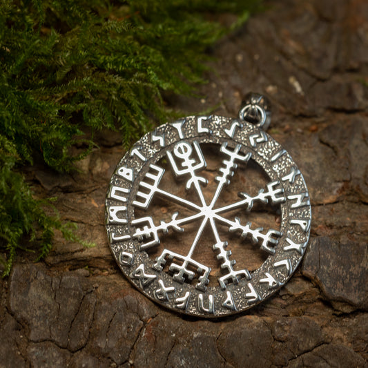 Et rundt Vejviser Vedhæng med Runer Stål vedhæng ligger på en struktureret barkflade ved siden af grønt mos. Vedhænget har indviklede runesymboler fra Futhark-løberen langs den ydre kant og et centralt design, der minder om et Vegvísir- eller kompashjul.