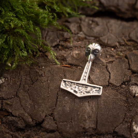 En lille, indviklet designet metalskulptur kaldet Thors Hammer Yilu Vedhæng 925s Sølv, med detaljerede raderinger, ligger på en struktureret, bark-lignende overflade. Mos er synligt i øverste venstre hjørne af billedet, hvilket vækker en følelse af vikingetiden og nordisk mytologi.