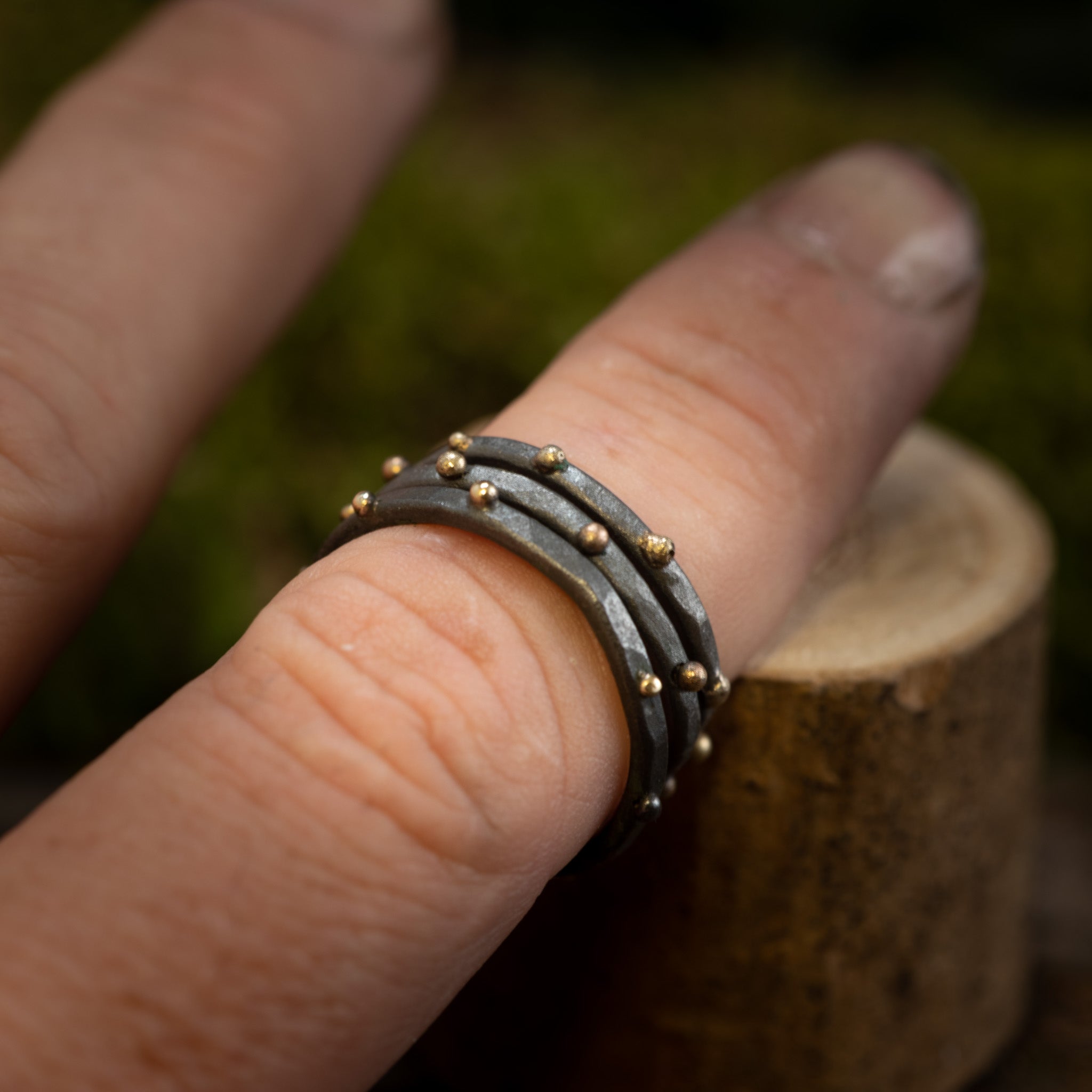 Nærbillede af en hånd, der viser en stak af tre hamrede 925 Sterling sølv ringe med små gyldne perler, fra Fingerring - Eryn Design - Eni kollektionen. De stabelbare ringe, som bæres på langfingeren, har et elegant dansk design, med en let sløret baggrund, der gør opmærksom på smykkets indviklede detaljer.