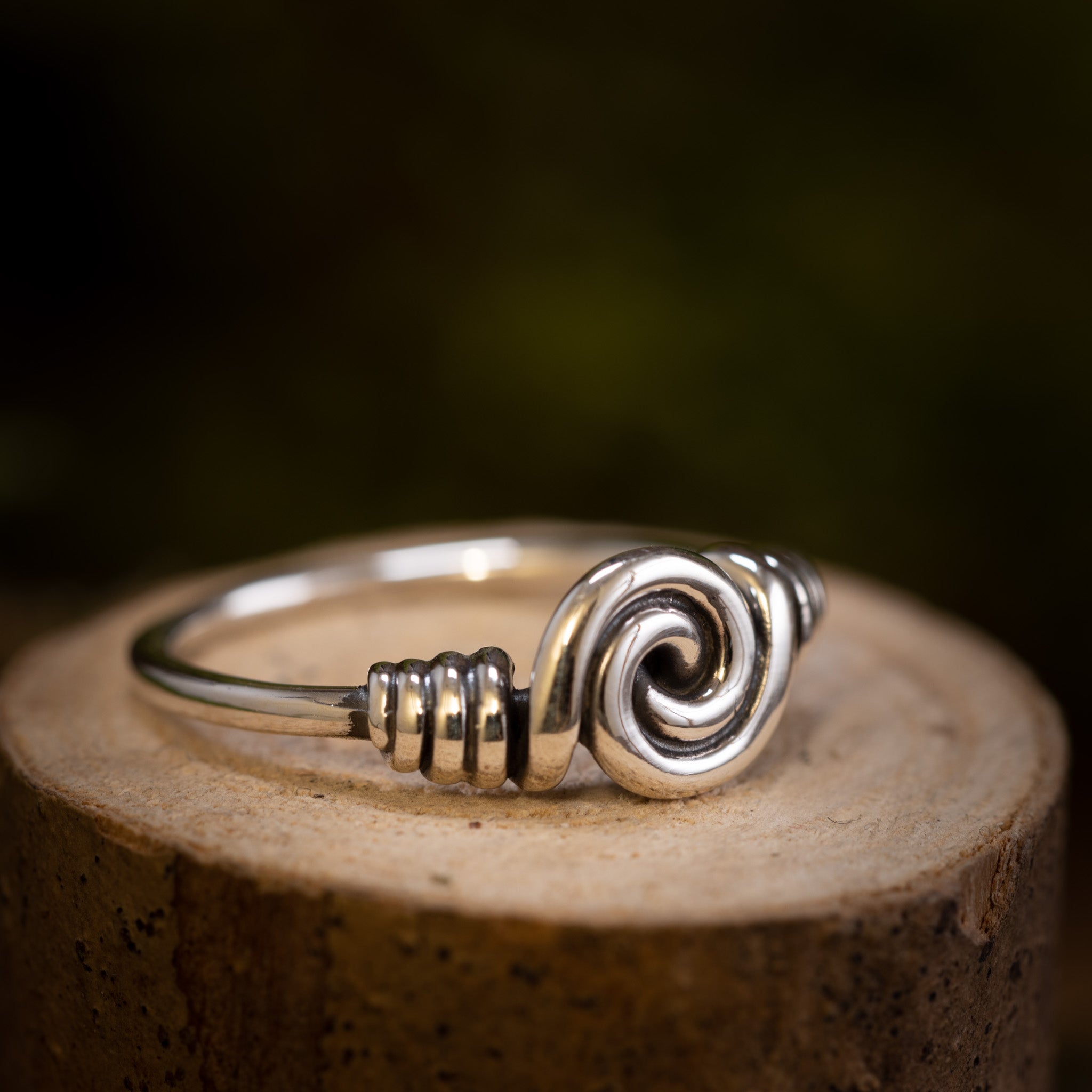 Et nærbillede af Sølvring Spiral 925s Sølv-ringen med et indviklet oprullet tråddesign i midten, der minder om Vikingerne håndværk. Ringen er placeret på en træoverflade med en mørk, sløret baggrund, der fremhæver ringens håndværk og skinnende, reflekterende egenskaber.