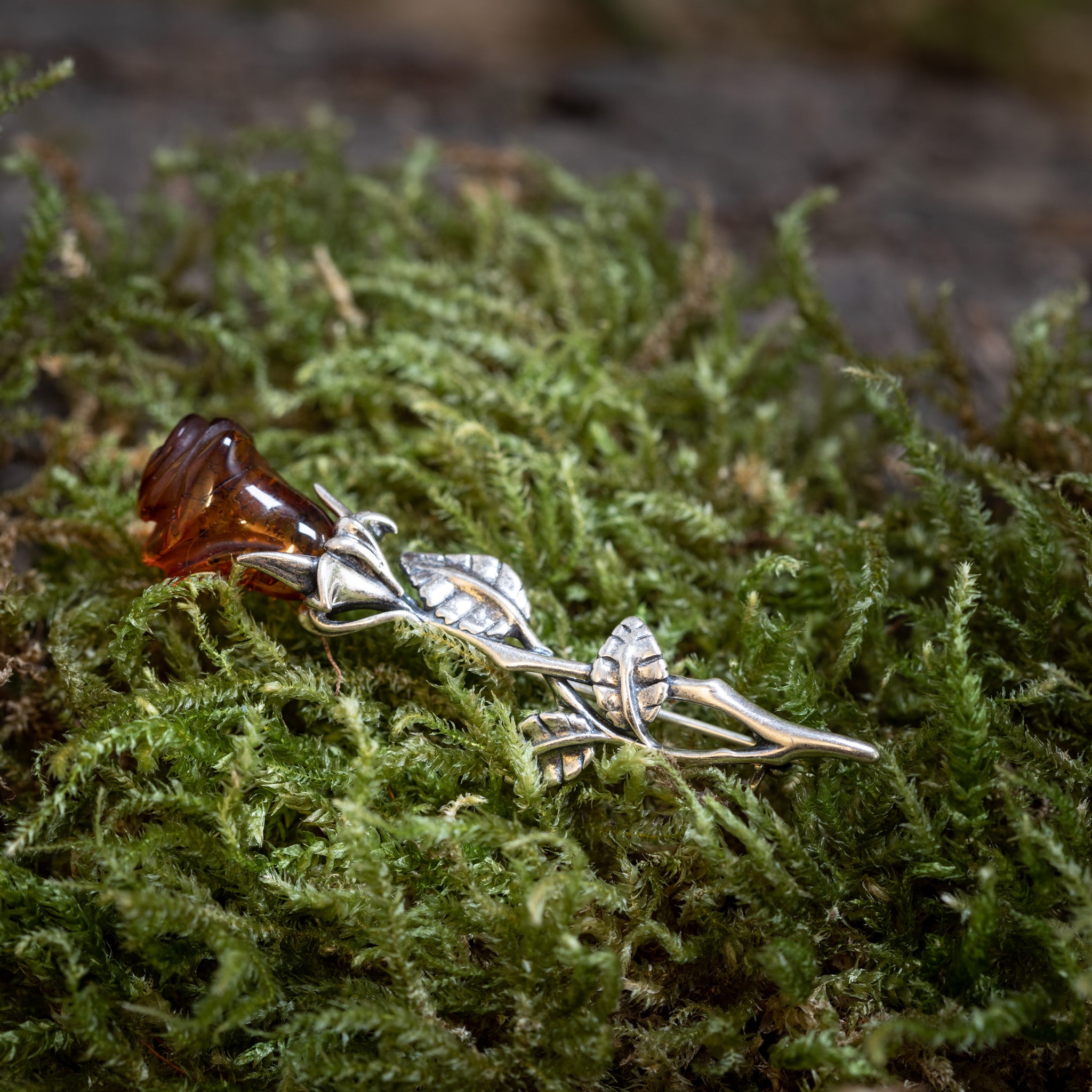 Broche Rose med Rav 925s Sølv, en sølvfarvet broche formet som en stilk med blade, holder autentisk rav, der ligner en blomsterknop, og hviler på levende grønt mos. Det detaljerede håndværk af denne romantiske gave efterligner en naturlig rose.