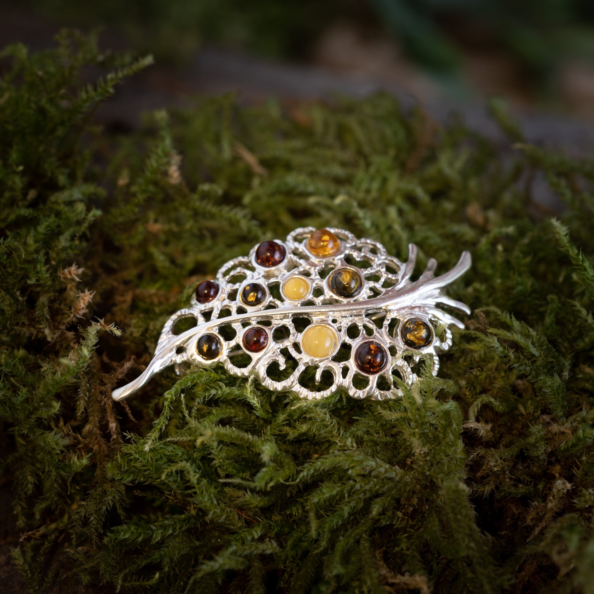 Et Broche Leaf med Rav 925s Sølv, formet som et blad med indviklet filigranarbejde, ligger puttet på et bed af frodigt grønt mos. Dette naturnær design har flere cirkulære ravdråber i ravfarver, der tilføjer farve og elegance til den detaljerede sølvbroche.