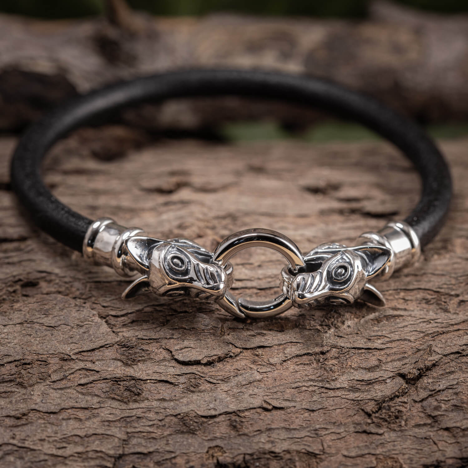 Armbånd Fenris Ulven 925s Sterling Sølv, med udsøgt Fenrisulv-hoveddesign i begge ender, er elegant spændt om et sort læderarmbånd. De to ulvehoveder, kyndigt udformet i Sterling Sølv, står over for hinanden, mens de griber om en central sølvring med munden. Armbåndet vises på en rustik træoverflade.