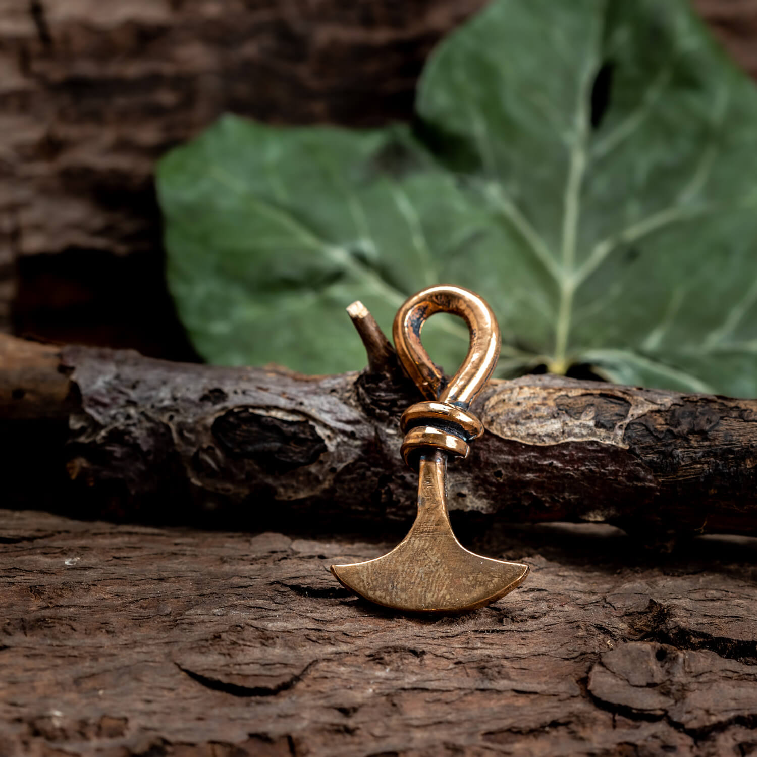 En lille rustik Thors Hammer Vedhæng Bronze ligger på en struktureret træoverflade. Et grønt blad, delvist sløret, er i baggrunden, hvilket giver en naturlig baggrund. Det økseformede vedhæng, der minder om Thors legendariske våben, har et indviklet buet håndtag med en ring nær toppen.