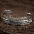 Bild in Galerie-Betrachter laden, Et nærbillede af et indviklet udformet sølvmanchetarmbånd. Armbånd Bangle Feather 925s Sterling Sølv har detaljerede, ætsede diagonale linjer, der efterligner teksturen af en kragefjer med en poleret finish. Armbåndet er lavet af 925s Sterling Sølv og er sat mod en rustik, brun træbaggrund.
