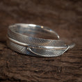 Bild in Galerie-Betrachter laden, Et nærbillede af Armbånd Bangle Feather 925s Sterling Sølv designet til at ligne en fuglefjer. Armbåndet har indviklede detaljer, med fine linjer, der efterligner teksturen af en ægte kragefjer. Det er placeret på en rustik træ baggrund.
