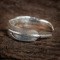 Bild in Galerie-Betrachter laden, "Armbånd Bangle Feather 925s Sterling Sølv" er et 925s sterlingsølv armbånd, der er indviklet designet til at ligne en detaljeret kragefjer, med den ene ende snoet rundt for at danne det åbne armbånd, smukt hvilende på en tekstureret træoverflade.
