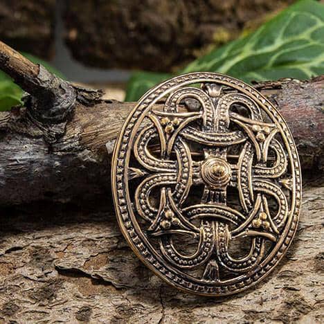 Broche Borrestil Bronze, en kompliceret designet broche i middelalderstil, der minder om autentisk vikingedesign, hviler på et stykke forvitret træ. Brochen har et udsmykket, symmetrisk mønster med sammenflettede detaljer og en central hævet knude, sat mod en rustik baggrund med grønt løv, der fanger essensen af Fæsted skatten.