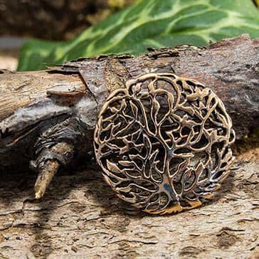 "Broche Yggdrasil Livets Træ Bronze", et indviklet udskåret rund bronzevedhæng, vises på en tekstureret træoverflade. Grøn vegetation i baggrunden tilføjer et naturligt præg til scenen.