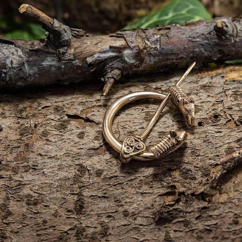 Broche Ringnål Stor Bronze, prydet med indviklede keltiske designs, hviler på et tekstureret stykke træ. Med trægrene og grønne blade i baggrunden fremkalder denne bronzering minder om vikingegenstande med sine detaljerede graveringer, der har hjertelignende former og spiraler.