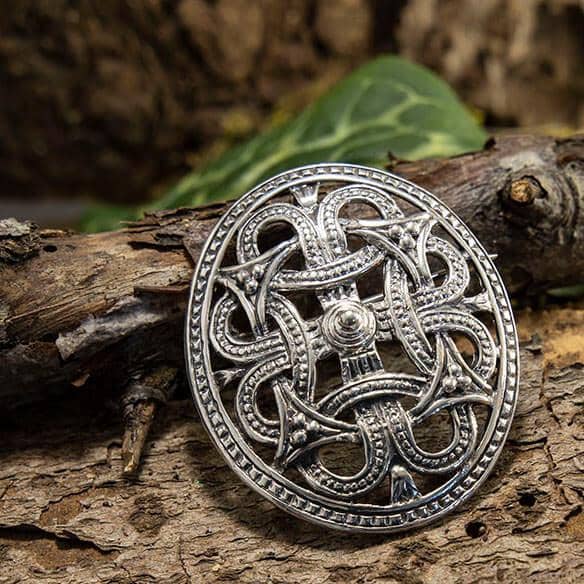 Broche Borrestil 925s Sterling Sølv, prydet med et delikat og komplekst keltisk knudedesign, er elegant udstillet på et stykke bark. Sammensætningen forstærkes af et grønt blad i baggrunden, der bidrager til den naturlige stemning. De omgivende rustikke omgivelser fremhæver brochens indviklede tekstur og overlegne håndværk, hvilket gør den til et lokkende stykke for enhver historieinteresseret.