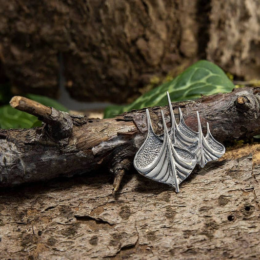 En detaljeret, bådformet metallisk broche i 925s Sterling sølv hviler på et groft stykke bark. Det indviklede design fanger lyset og viser dets håndværk. Grønt løv i den slørede baggrund tilføjer et naturligt touch til scenen.