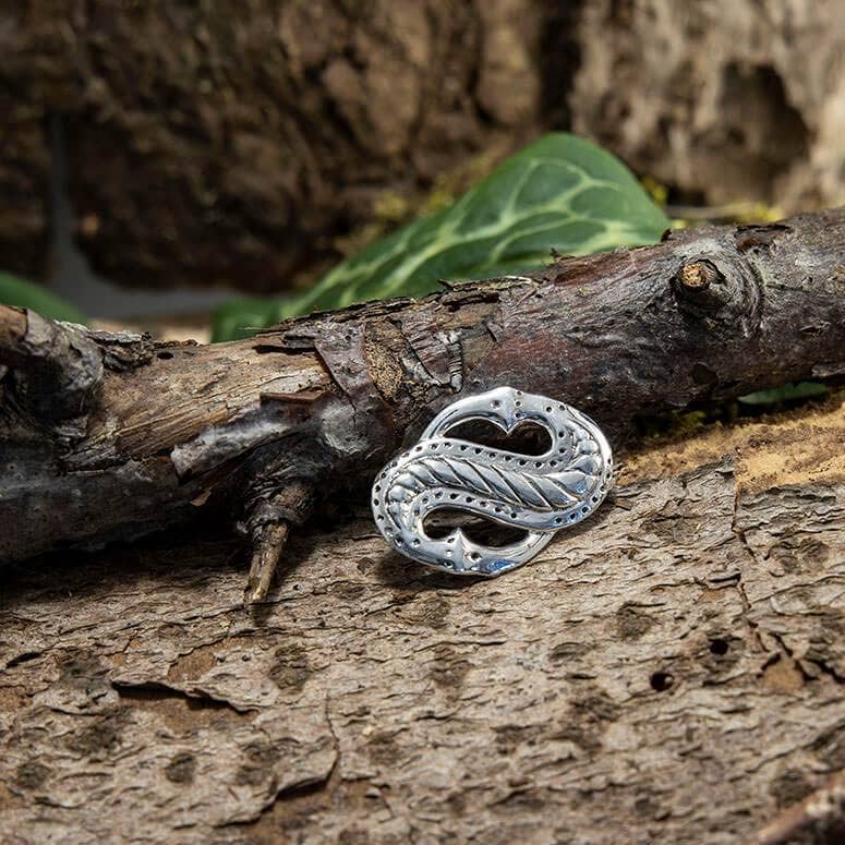 En detaljeret Broche Midgårdsormen 925s Sterling Sølv med en sammenflettet, tekstureret knude er placeret på en ru bark. Grønne blade giver en naturlig baggrund, der forstærker den rustikke og jordiske æstetik ved dette Vikingetiden-symbol.