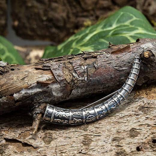 En Broche Guldhorn 925s Sterling Sølv, med symboler og indviklede designs, der minder om en vikingebroche, er viklet rundt om en mosklædt trægren. Baggrunden har store grønne blade og jordfarver, hvilket skaber en naturlig skovstemning i scenen.