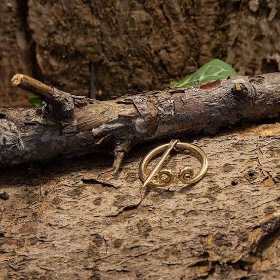 En Broche Ringnål Lille Bronze med et hvirvlende design ligger på den træagtige, teksturerede overflade af en trægren, der blander sig med de naturlige, jordiske omgivelser. Blade og bark tilføjer den rustikke stemning i denne scene, der minder om vikingetiden.