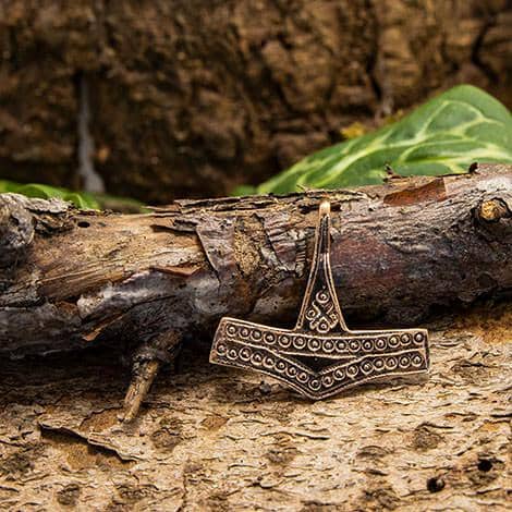 En Thors Hammer Romersdal Vedhæng Bronze, designet som museumskopi af Thors Hammer, ligger på en ru, forvitret træstamme med en baggrund af grønne blade og struktureret bark. Vedhænget er prydet med indviklede prikmønstre.