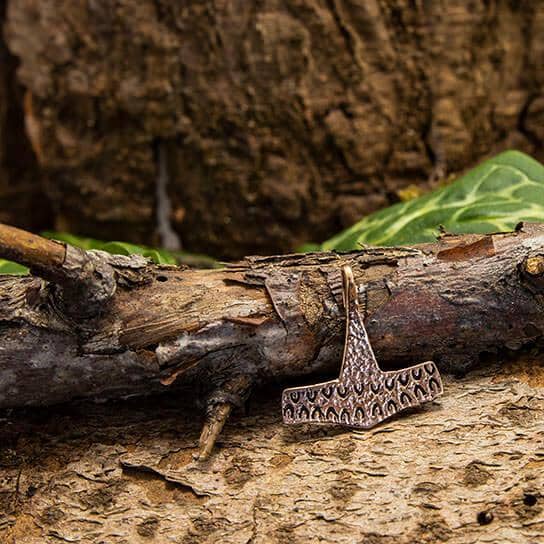 Et rustikt tekstureret Thors Hammer Strandby Vedhæng Bronzevedhæng, der minder om vores vikingeforfædre, sidder på en forvitret trægren med grønne blade delvist synlige i baggrunden og blander natur og håndværk.