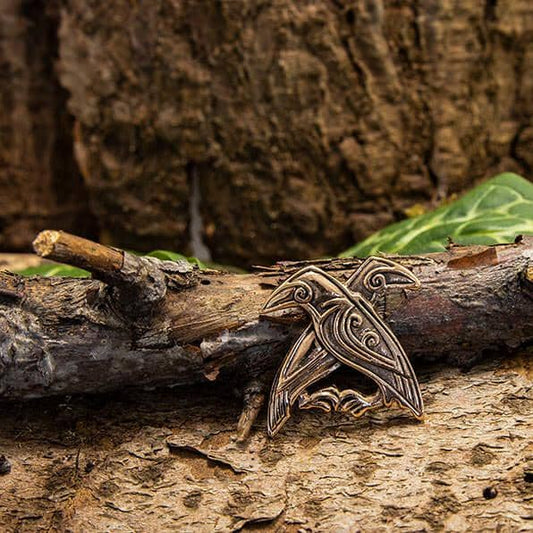 En Hugin og Munin Stor Vedhæng Broche af bronzemetal med to indviklede fugle er placeret på et stykke træ. Baggrunden viser ru bark og grønne blade, hvilket skaber et naturligt og rustikt miljø. Brochen har en detaljeret, kunstnerisk graveringsstil, der minder om Odins ravne, Hugin og Munin.