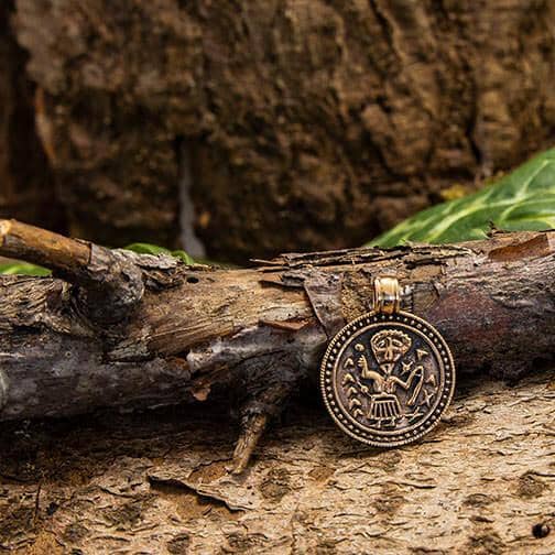 En rund amulet Frigg Vedhæng bronze med indviklede graveringer hviler på en struktureret træoverflade, på en baggrund af træbark og grønne blade. Designet på vedhænget har detaljerede og symmetriske mønstre, hvilket tilføjer dens rustikke charme.