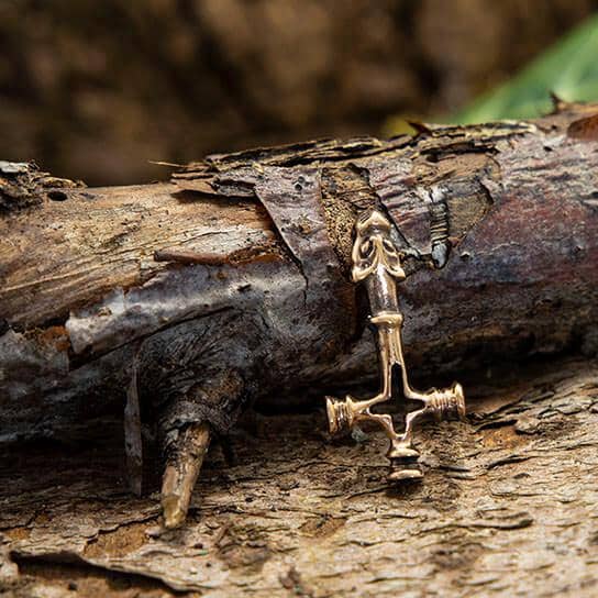 Et nærbillede af Islandsk Kors Vedhæng Bronze, et lille vedhæng, der er indviklet formet som et ø-kors, placeret på struktureret og forvitret træbark. Korset har detaljerede designs og små figurer i krydset. Den ru tekstur af barken i baggrunden tilføjer en rustik følelse til billedet.