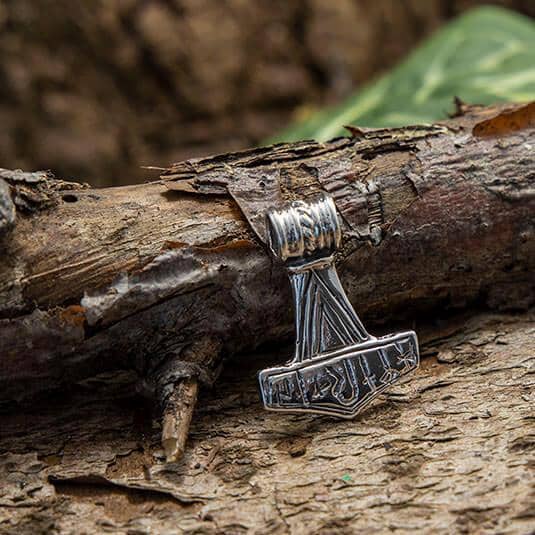 En slank Thors Hammer Vedhæng Celtic 925s Sølv, inspireret af Mjolnir fra vikingetiden, hviler mod en forvitret trægren med skovbunden i baggrunden. Ægte sølv vedhænget kan prale af indviklede graveringer, hvilket tilføjer dets detaljerede udseende.