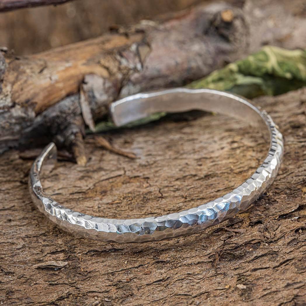 Armbånd Bangle Sya 925s Sterling Sølv hviler på en rustik træoverflade med kviste og blade i baggrunden. Det åbne armbånd er lavet af 925s Sterling Sølv og har en hamret finish, hvilket giver det et robust, men elegant udseende.