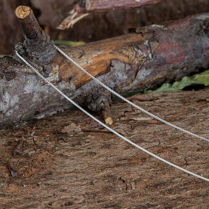 Halskæde Venezia 925s Sølv 0,8 mm drapererer delikat over en robust trægren, hvilende mod en struktureret, jordagtig baggrund. De raffinerede smykker står i smuk kontrast til de naturlige omgivelser.