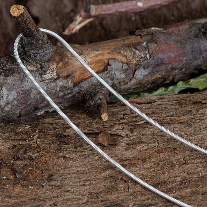 En metalkabelsav ligger på en ru træoverflade, sandsynligvis til brug i udendørs- eller overlevelsessituationer. Saven er oprullet med to løkkehåndtag i hver ende, designet til let greb. Blandt stykkerne af træbark og blade kan man se et glimt af en Halskæde Slangekæde 925s Sølv 2mm, der tilføjer et uventet strejf af elegance.