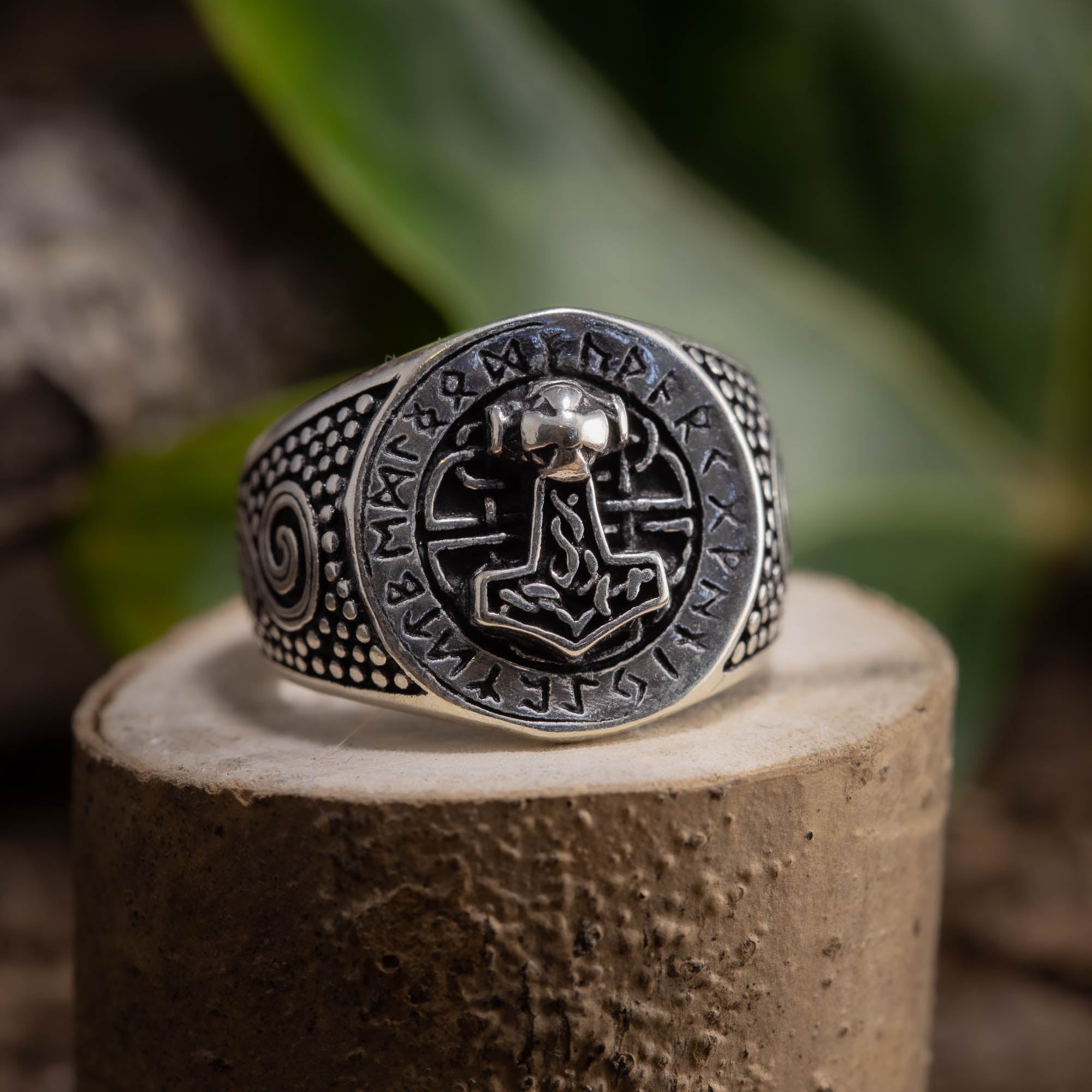 Sølvring Thors Hammer 925s Sølv-ringen viser stolt et detaljeret Mjölnir (Thors Hammer)-emblem i midten, omgivet af indviklede runer og mønstre. 925 Sterling sølv ringen hviler på en struktureret stensokkel med en sløret grøn bladagtig baggrund.