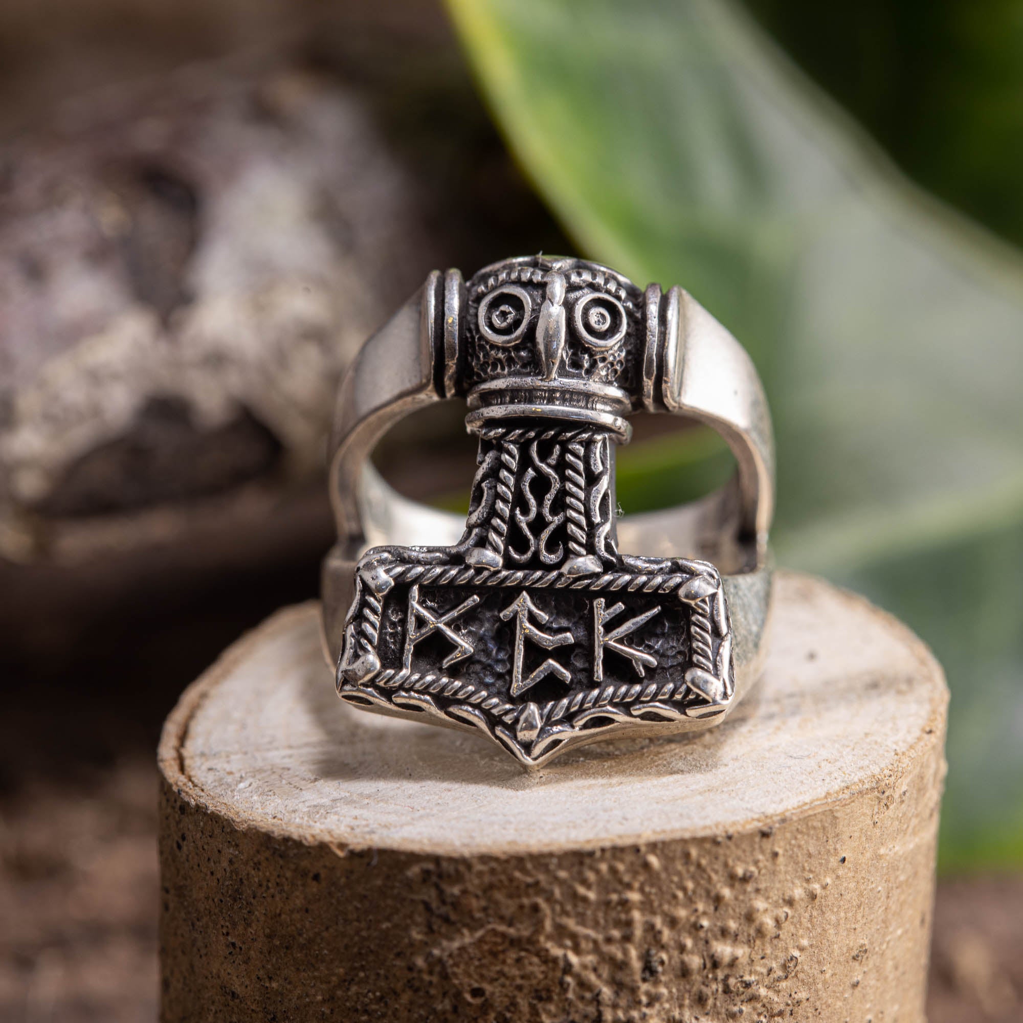 Sølvring Thors Hammer 925s Sølv, en ring i sterling sølv med indviklede nordiske designs, sidder på en træsokkel. Thors-hammeren fungerer som midtpunktet, prydet med runer og detaljerede udskæringer. Dette udsøgte stykke, fås i forskellige ringstørrelser, er sat mod grønt løv og en sløret naturlig indstilling.