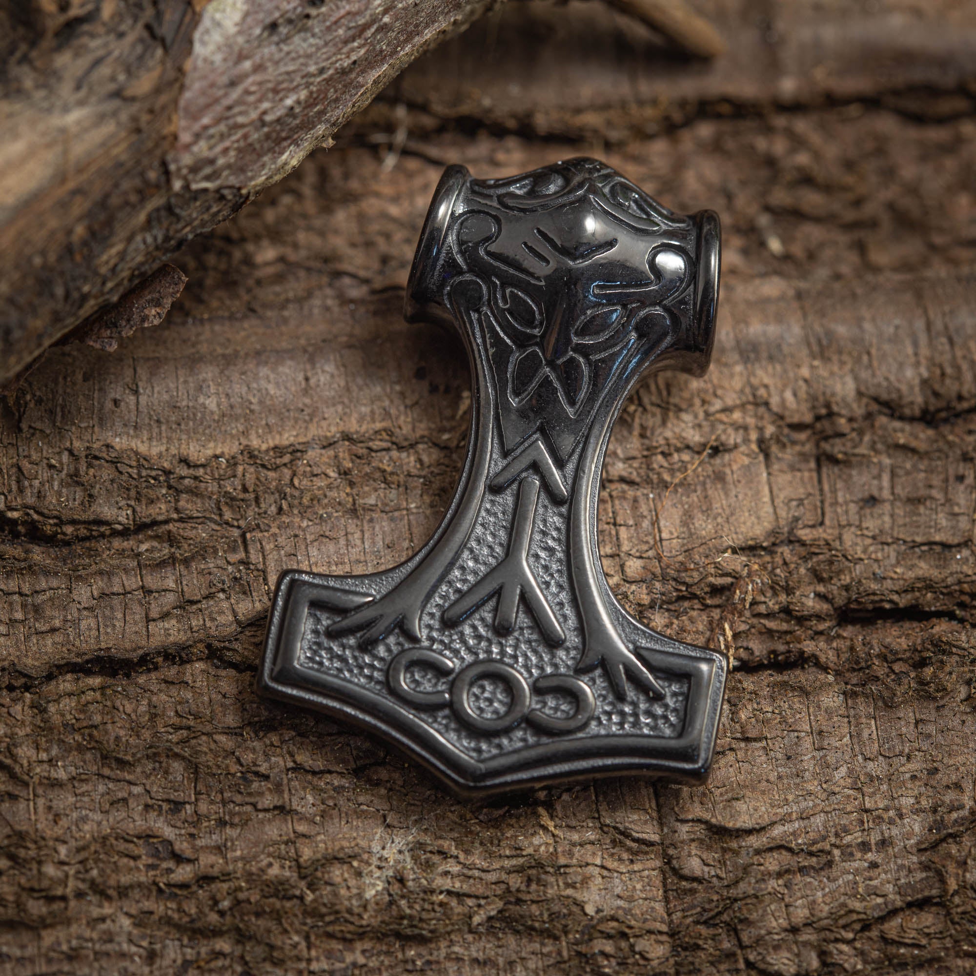 En Thors Hammer Vedhæng Sort Stål, lavet i sort metal og ligner Mjölnir, ligger på en struktureret, rustik træoverflade. Dette stykke fra BlaX Steel har indviklede indgraverede nordiske designs, der inkarnerer elegancen og styrken, der er synonymt med fine stålsmykker.