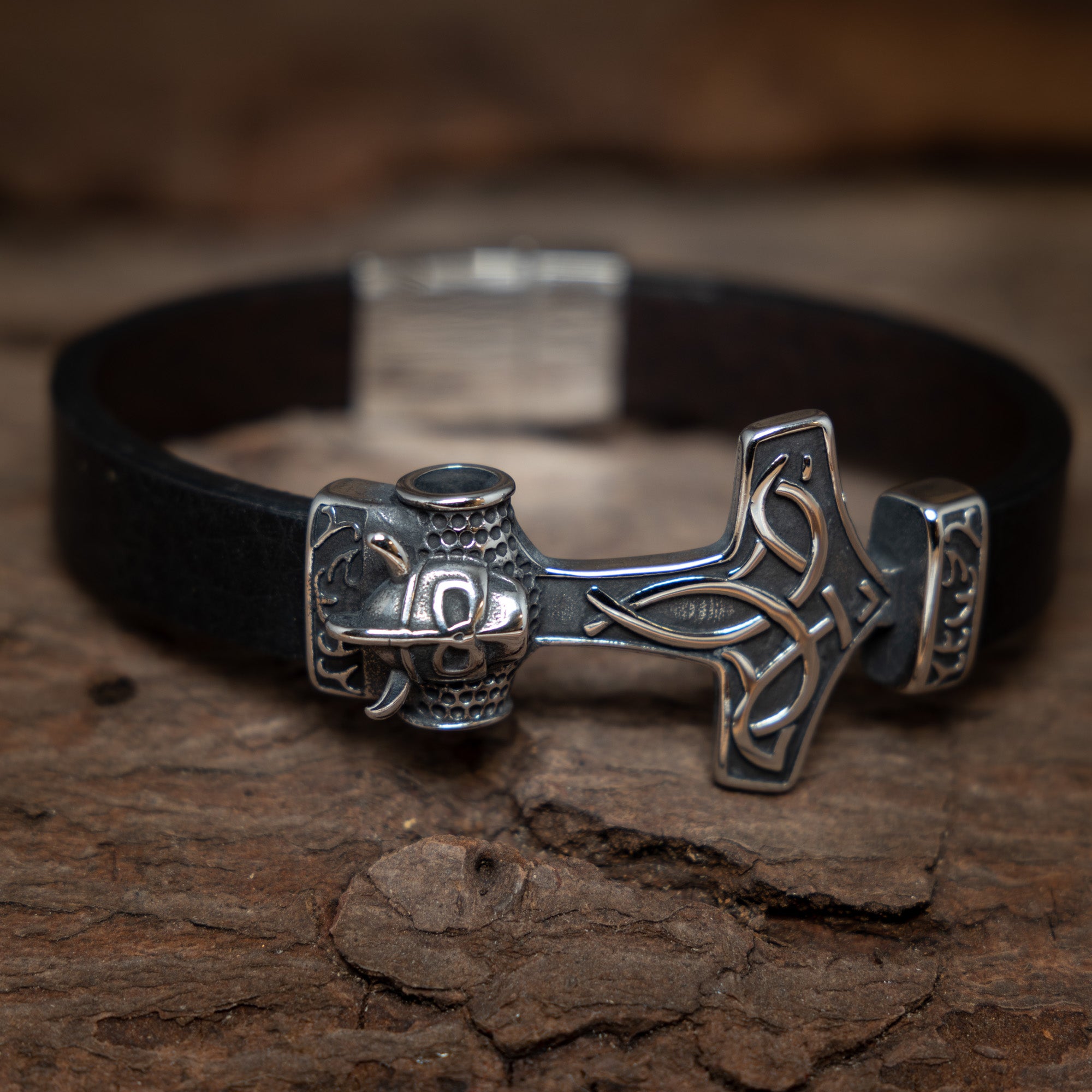 "Armbånd Thorshammer m. Hjelm Stål," med et sort læder-armbånd prydet med et sølv Thors hammerformet vedhæng med indviklede detaljer med nordisk-inspirerede designs, er placeret på en tekstureret træoverflade. Låsen er sammensat af børstet sølvmetal, der komplementerer armbåndets robuste, men raffinerede udseende.