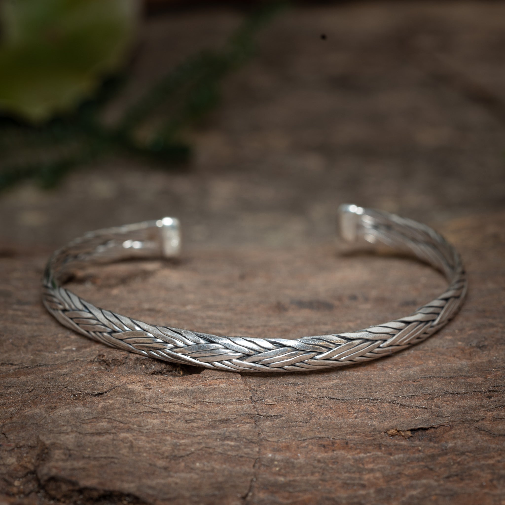 Armbånd Bangle Sield 925s Sterling Sølv, et fantastisk sølvflettet manchetarmbånd lavet af 925s Sterling Sølv, hviler på en rustik træoverflade med blødt, grønt løv i den slørede baggrund. Justerbart armbåndet har et indviklet vævet mønster og et åbent design.