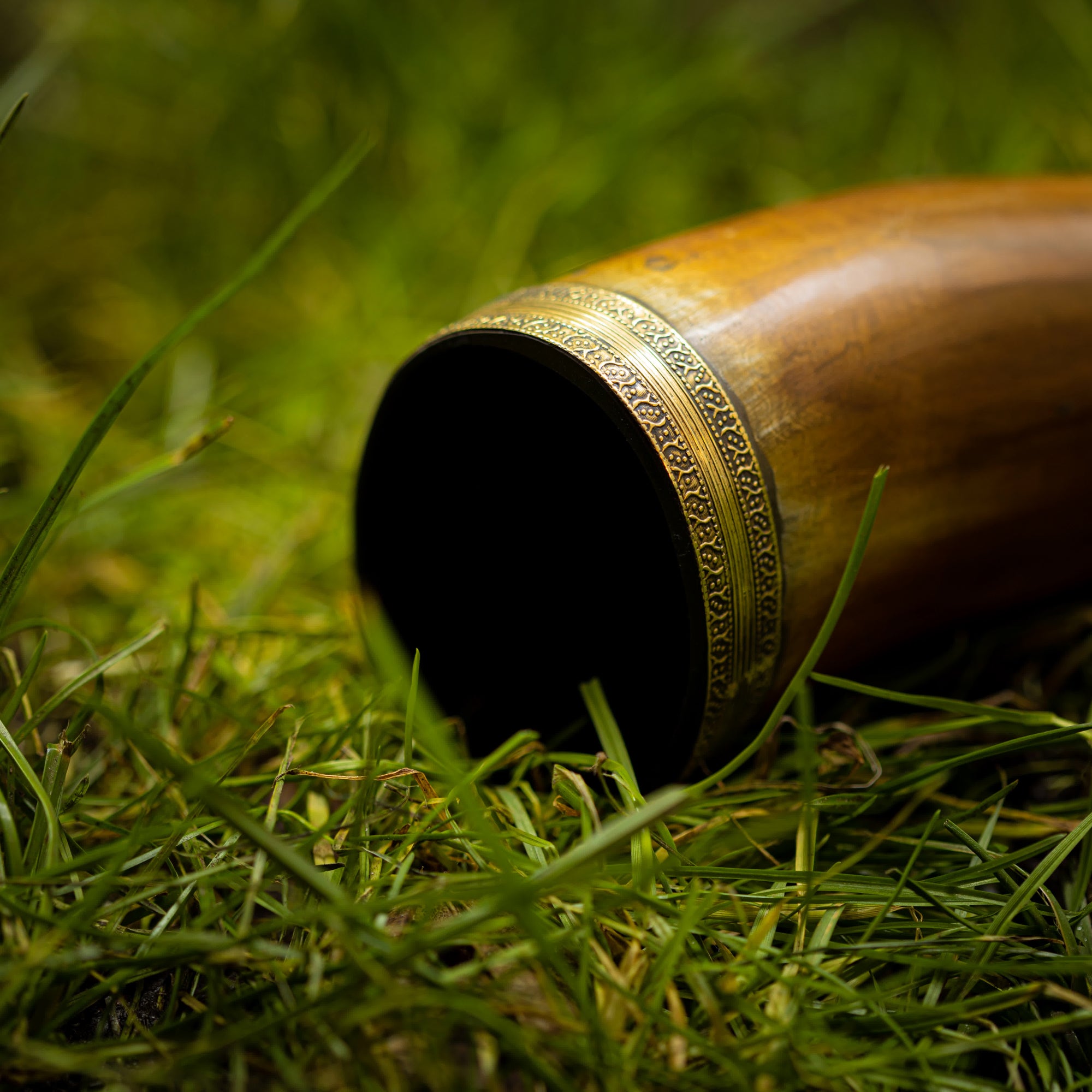 En smukt udformet Drikkehorn Devine med udsmykkede metaldetaljer på kanten ligger på grønt græs. Dette cylindriske drikkehorn af træ indsnævres mod den ende, der ikke er synlig på billedet. Det indviklede metalværk har mønstrede graveringer, der minder om dem, man fandt på vikingemarkeder.