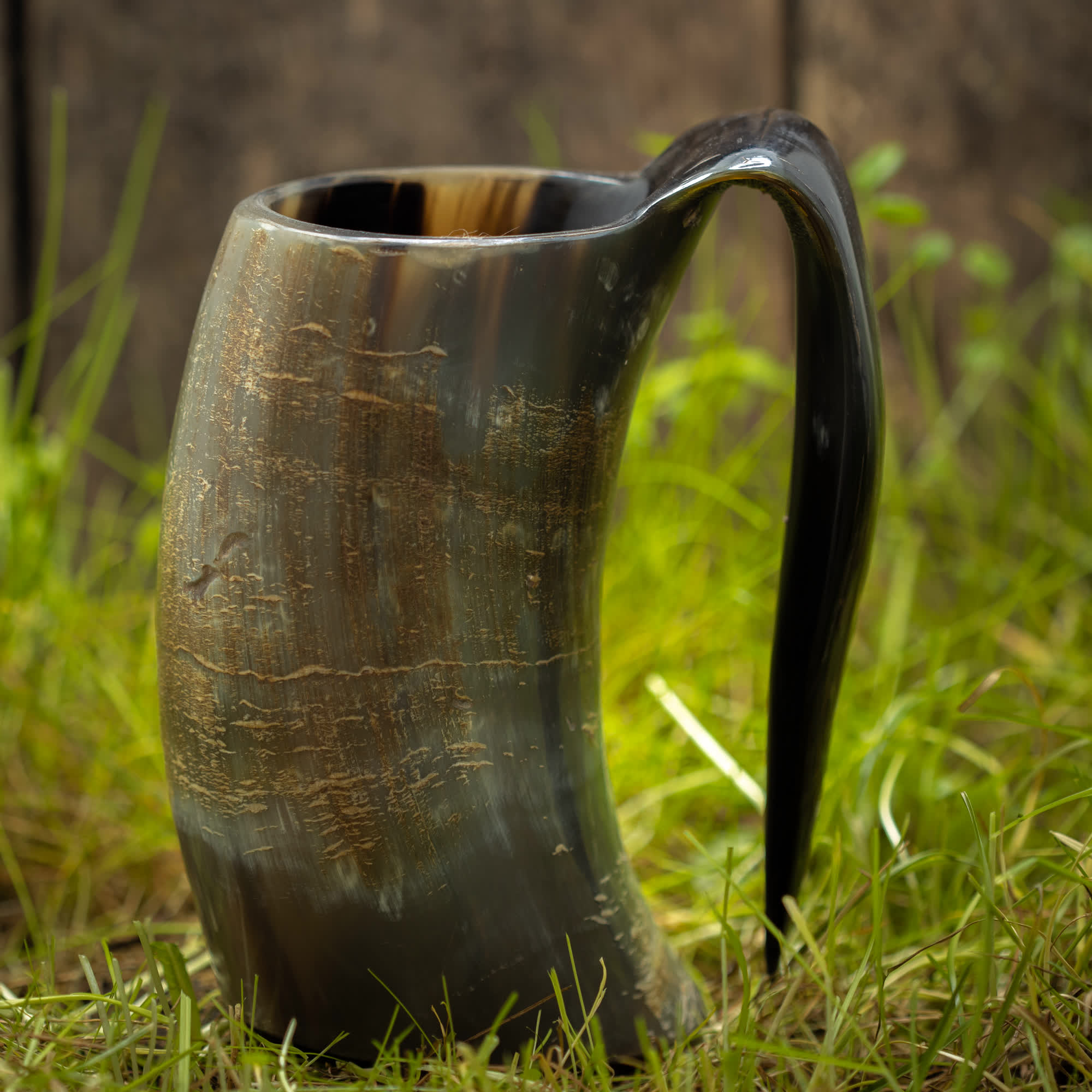 Et rustikt Vikingekrus Nature Horn (600-800ml) med et håndtag er placeret på grønt græs. Krus har et poleret, marmoreret mønster med nuancer af brun, sort og beige, der smukt reflekterer lyset. Baggrunden er let sløret og antyder en træoverflade.