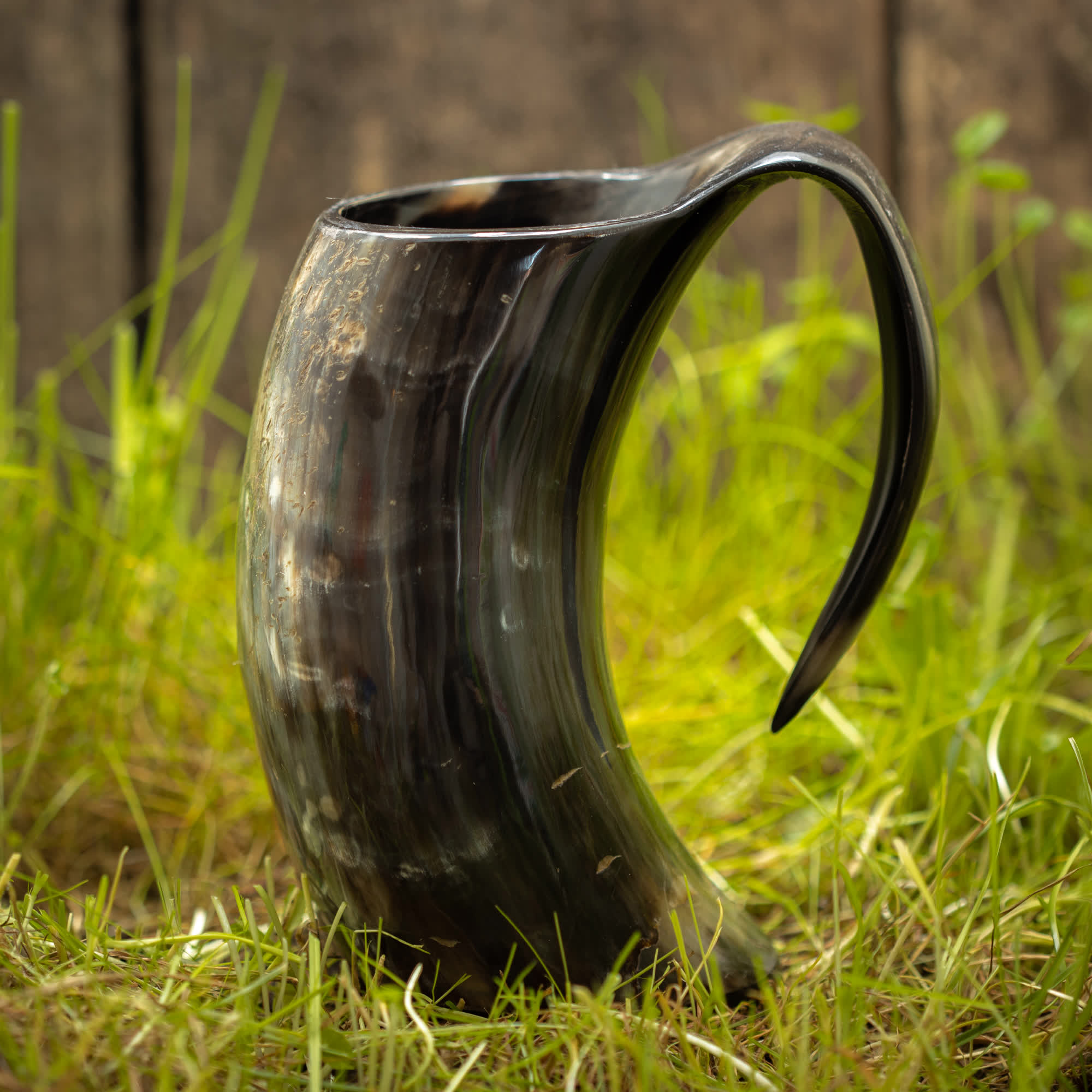 Et poleret Vikingekrus Nature Horn (600-800ml) med en mørk finish og buet håndtag står oprejst på en plet af grønt græs. Baggrunden er let sløret, med en træoverflade.