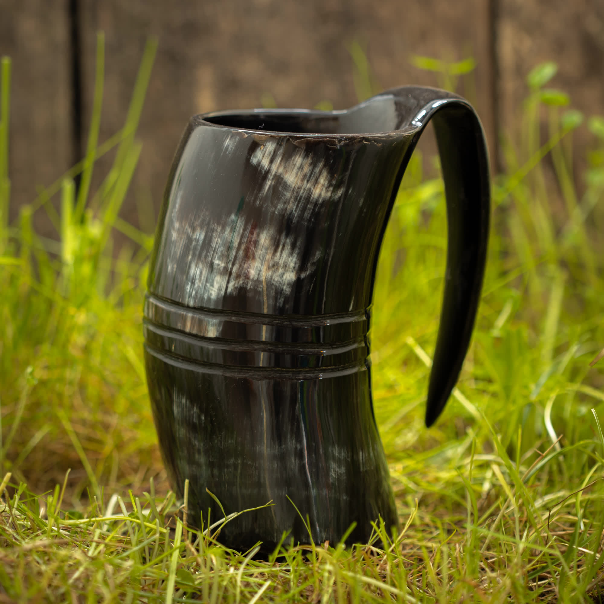 Et buet, sort Vikingekrus Helleristning Horn (600-800ml) sidder på en græsklædt overflade, med en træbaggrund i det fjerne. Kruset har et hank og har naturlige lyse og mørke striber, hvilket tilføjer dets rustikke udseende. Denne vikingekrus fremkalder en følelse af gammelt håndværk.