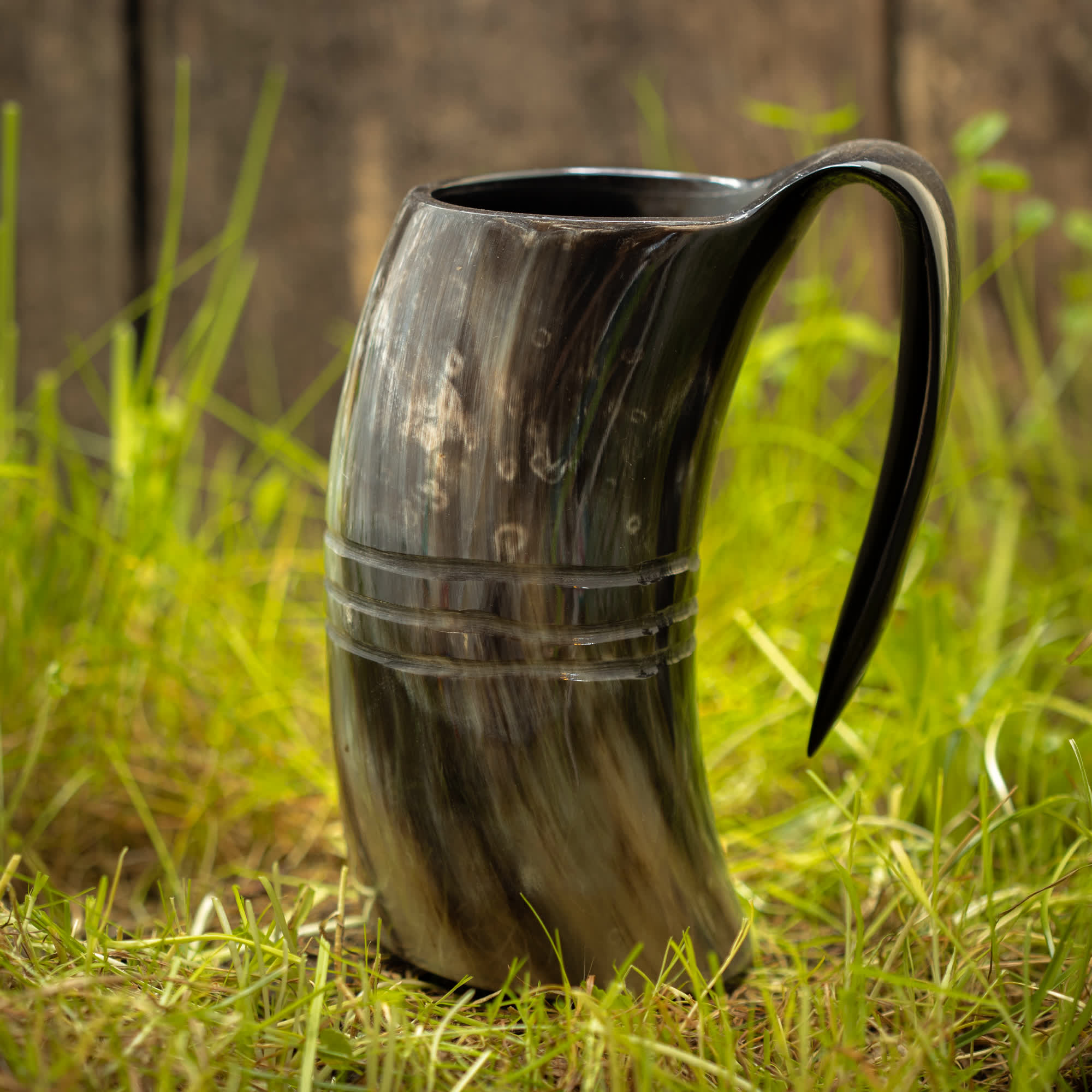 Et rustikt, håndlavet Vikingekrus Helleristning Horn (600-800ml) med et håndtag sidder på græs mod en træbaggrund. Viking Krus har en blank finish, der viser naturlige hornmønstre i nuancer af sort, brun og creme, med to indgraverede bånd rundt om midten for ekstra detaljer.