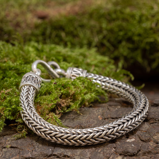 Et nærbillede af en Armbånd ByKila Sildeflet 5mm 925s Sølv liggende på en mosbeklædt overflade. Armbåndet, der er lavet af 925s Sterling Sølv, har et detaljeret flettet design med udsmykkede endestykker og en kroglås. Det naturlige mos og teksturerede baggrund står i kontrast til det polerede metal.