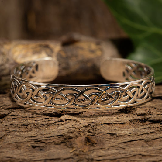 Armbånd Bangle Elvira 925s Sterling Sølv, med et indviklet keltisk knudedesign, hviler på en tekstureret træoverflade med en sløret grøn baggrund. Dette justerbare 925s Sterling Sølv-stykke fremhæver det detaljerede vævemønster og viser dets håndværk og elegance, der minder om vikingetiden smykker.