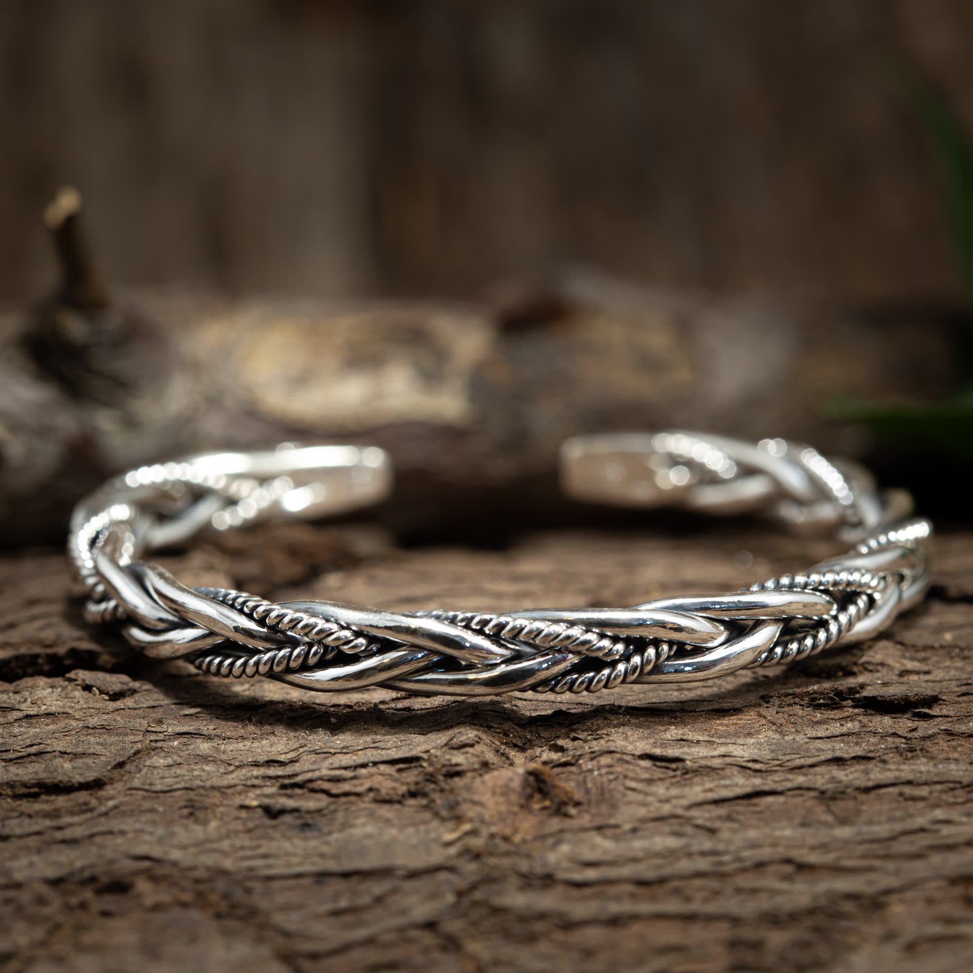 Et nærbillede af et indviklet designet Armbånd Bangle Laced 925s Sterling Sølv med et flettet mønster, placeret på en tekstureret træoverflade. Vævet armbånd har både glatte og reb-lignende elementer vævet sammen, hvilket viser detaljeret håndværk.