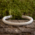 Bild in Galerie-Betrachter laden, Et Armbånd Bangle Empor 925s Sterling Sølv hviler på et stykke bark, med grønt mos sløret i baggrunden. Armbåndet har et indviklet, vævet design.
