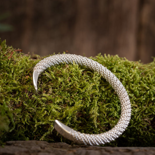 Et Armbånd Bangle Empor 925s Sterling Sølv vises på en blød, grøn mosbeklædt overflade med en sløret naturlig brun baggrund. Armbåndet er formet i en blid bue, der viser dets komplicerede design og håndværk.