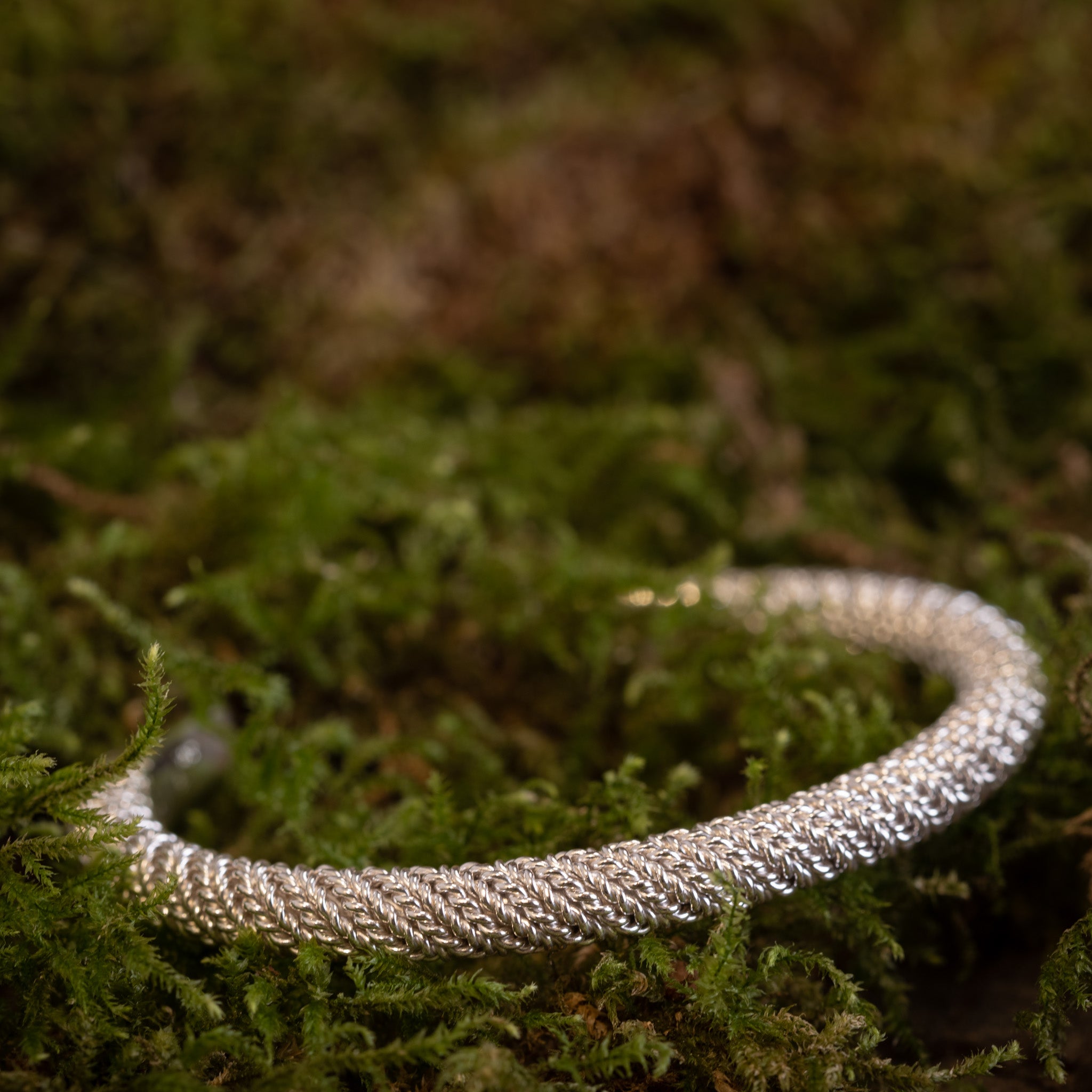 Et Armbånd Armbånd Empor 925s Sterling Sølv, et fantastisk sølvflettet armbånd, ligger på en seng af levende grønt mos. Det indviklede design står i kontrast til den naturlige, frodige baggrund, hvilket understreger dets detaljerede håndværk.