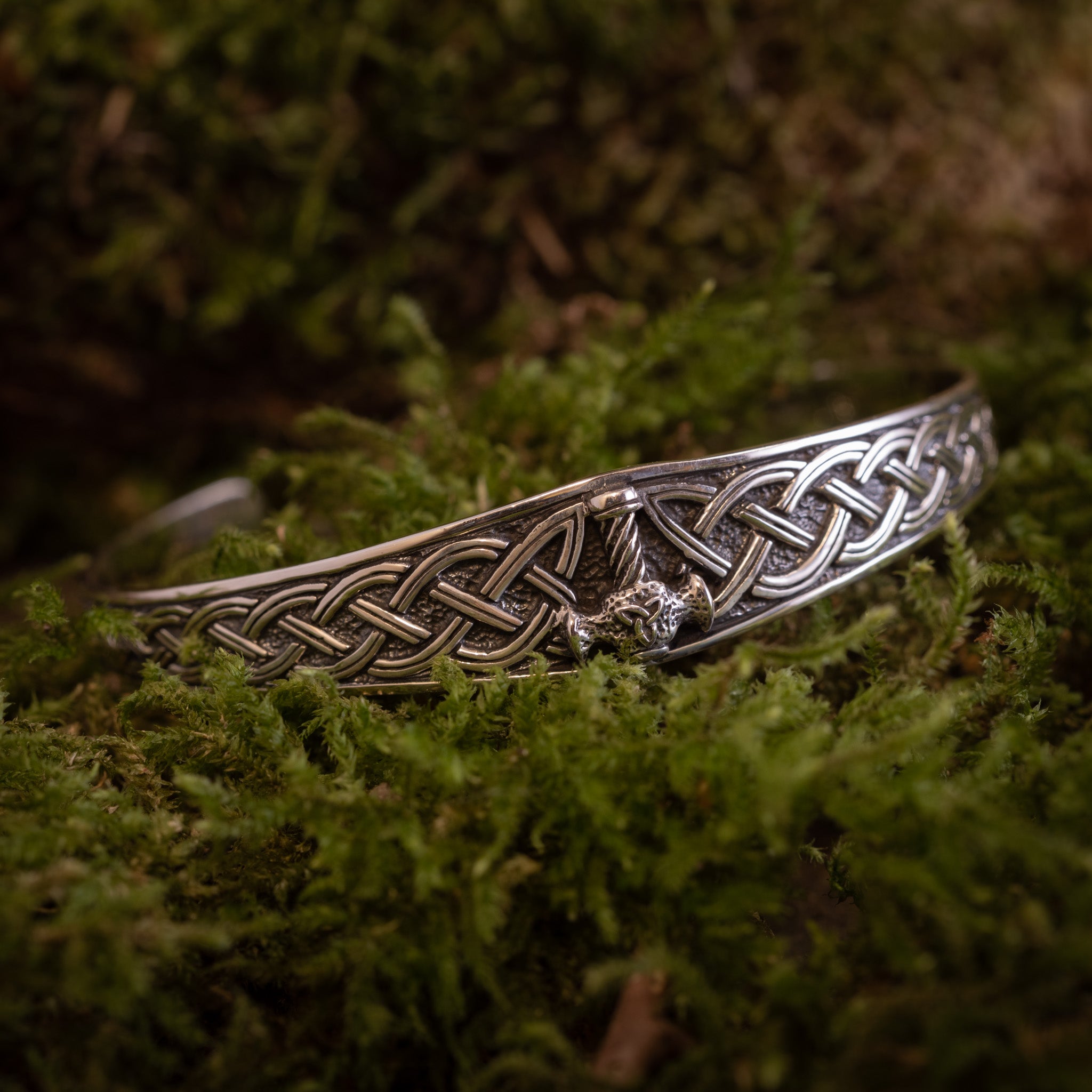 Et detaljeret Armbånd Armbånd Mjølnir lavet af 925'erne Sterling Sølv, med indviklede keltiske knudemønstre og et lille dragehoved i midten, sat mod en baggrund af frodigt grønt mos.