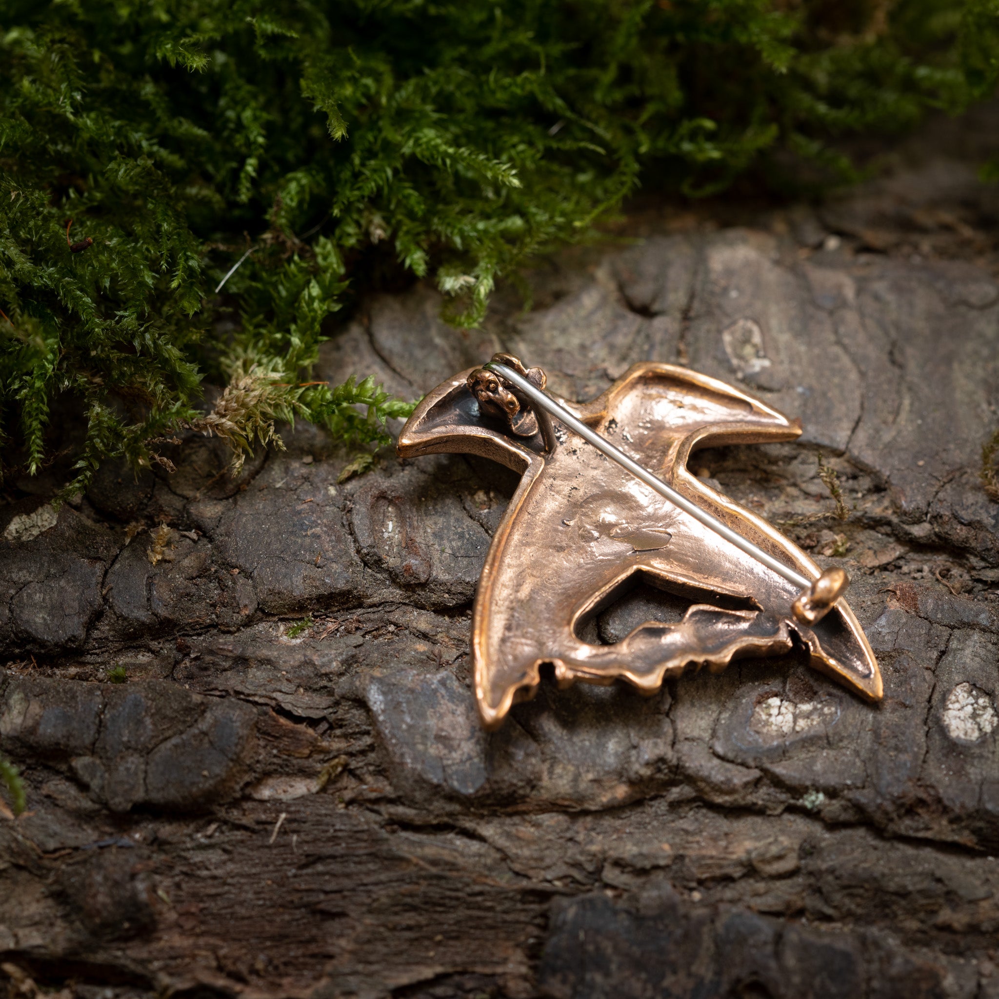 En lille, indviklet designet Broche Hugin og Munin Bronze i form af en fugl hviler på en struktureret træbarkoverflade med grønt og mos i baggrunden.