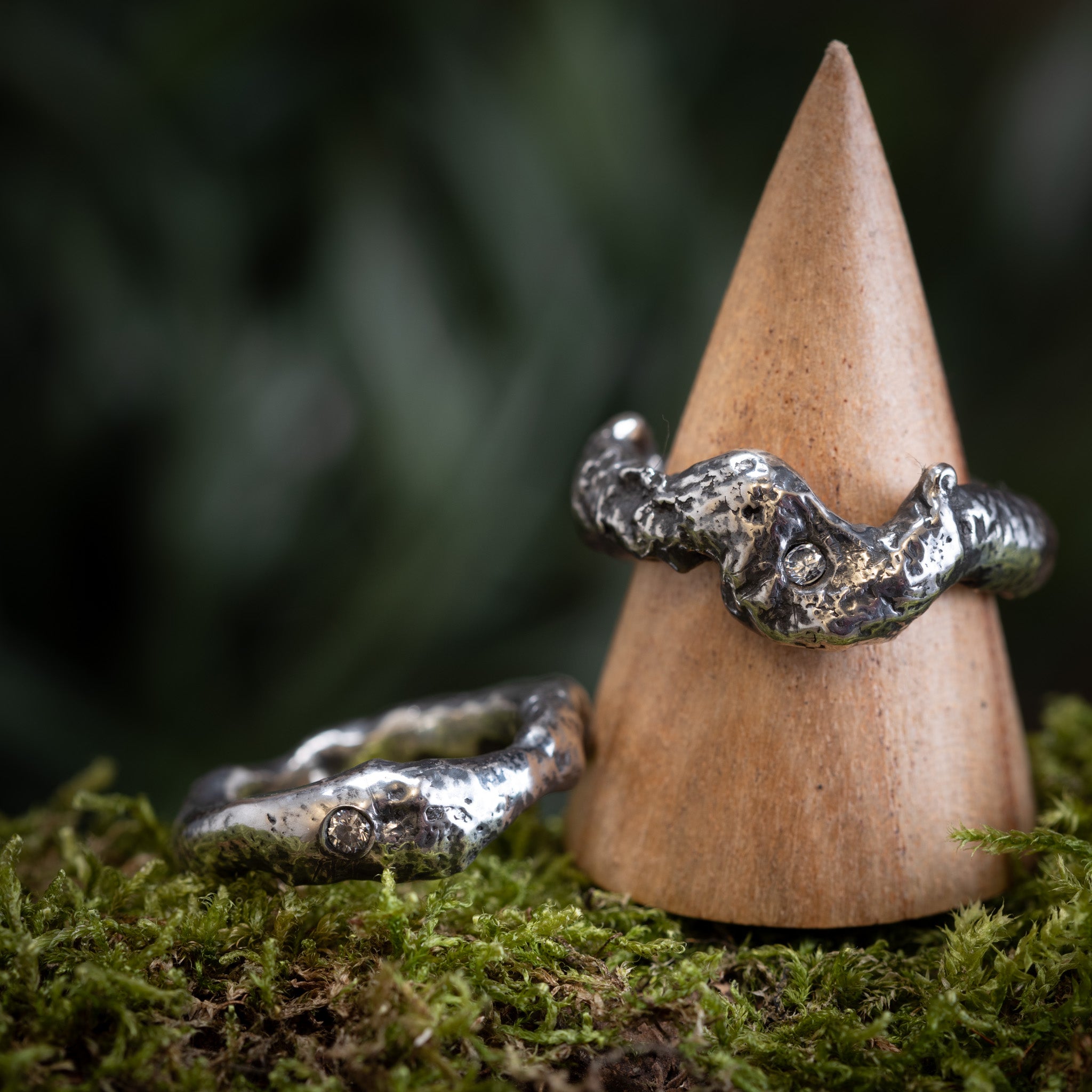 To "Fingerring - Eryn Design - Trial by Fire" stykker i 925s Sterling sølv vises på baggrund af grønt mos. Den ene ring hviler på jorden, og den anden er balanceret oprejst på et trækeglestativ. Begge håndlavede smykker har en ru, struktureret overflade og har små, indlejrede ædelstene.