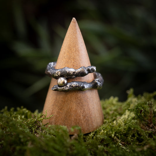 Fingerringen - Eryn Design - Treasure, en rustik ring fremstillet ved hjælp af Molten Silver-metoden og udsmykket med et tekstureret, naturinspireret design og en lille rund gylden perle, vises på en kegleformet træstativ. Stativet er sat på en bund af mos mod en sløret grøn baggrund, der fremhæver Eryn Designs udsøgte kunstnerskab.